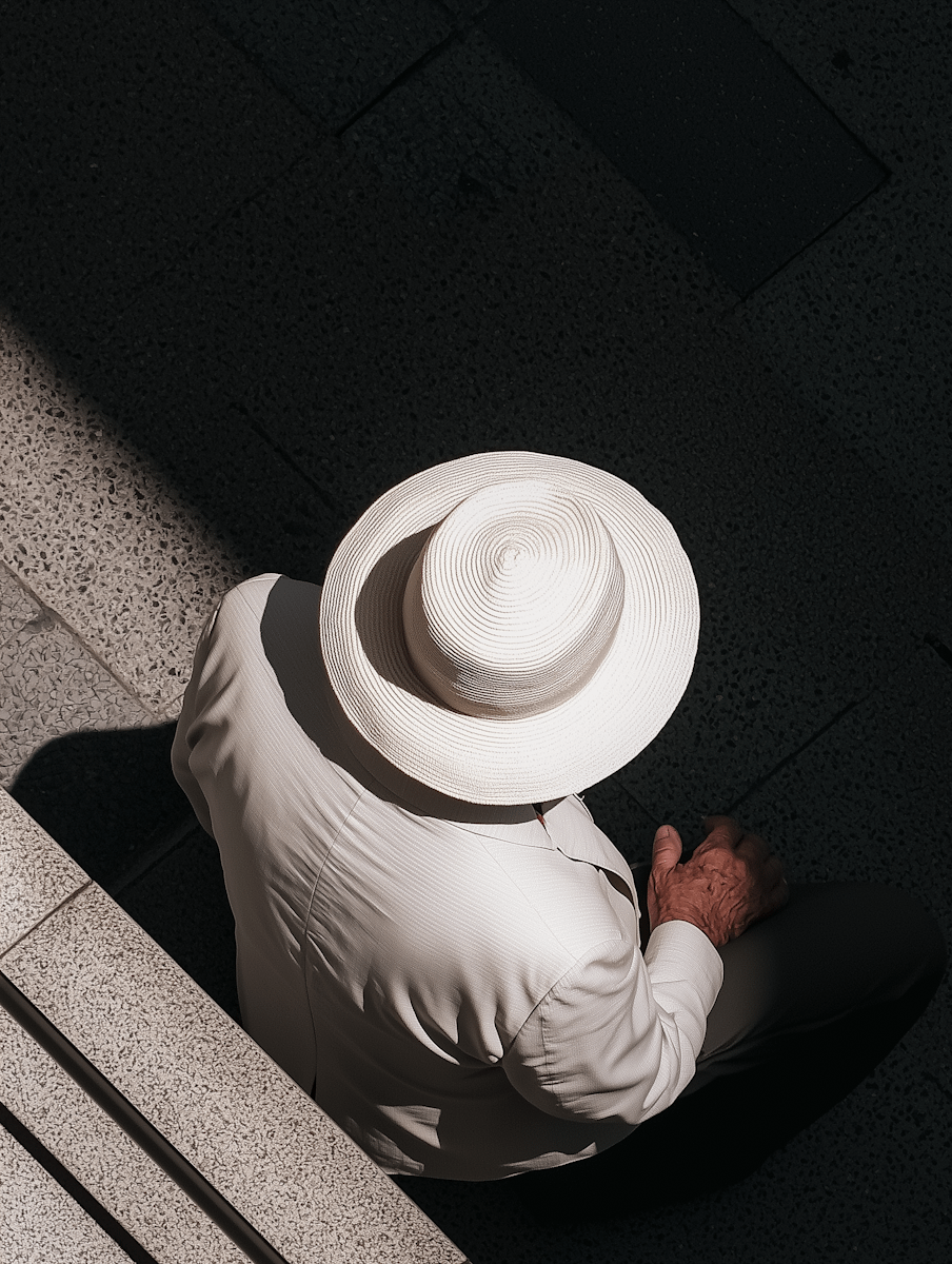 Top-down view of an elderly person in a white suit and hat sitting on a stone bench, with dramatic lighting and shadow contrast.