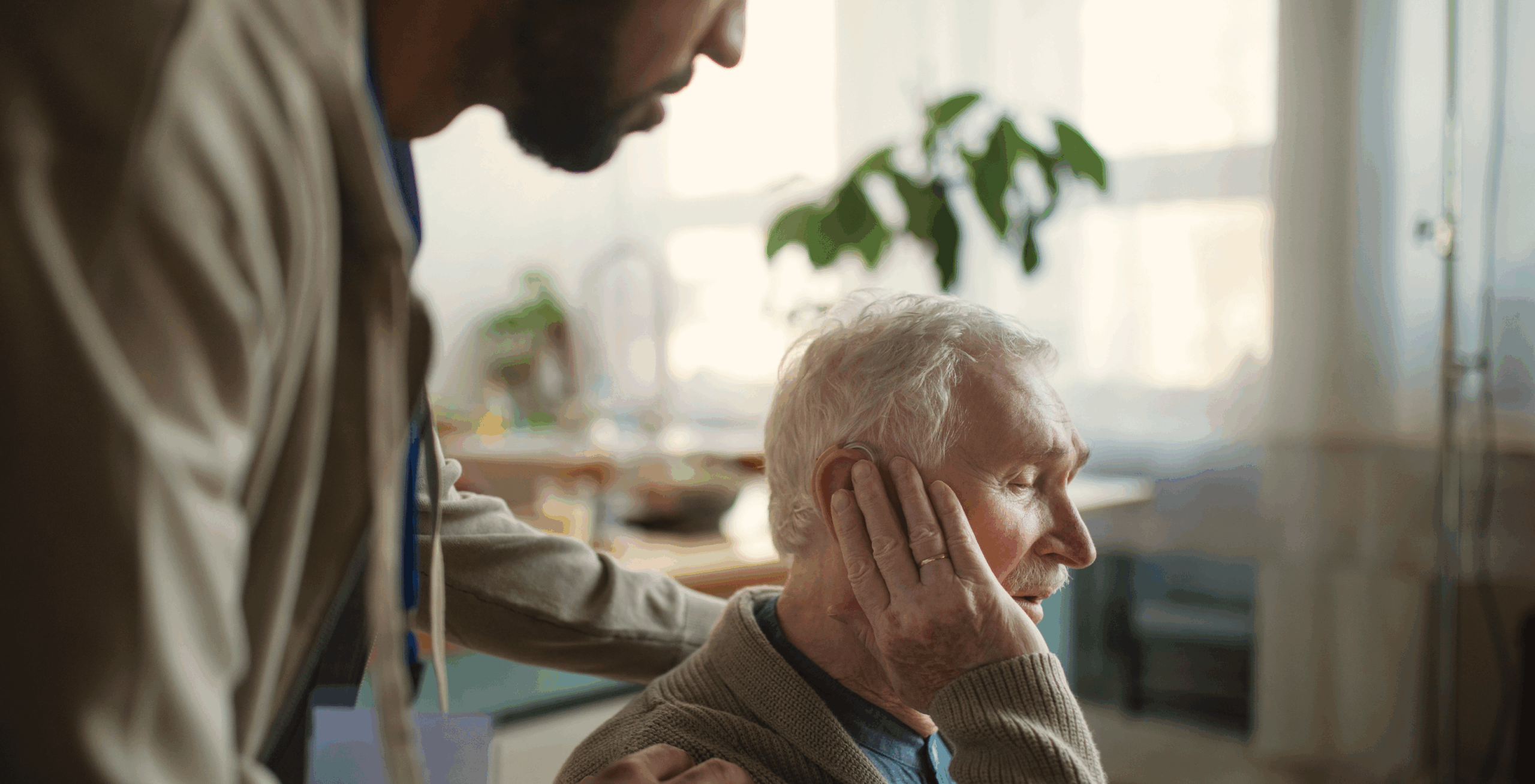 An old man wearing a wedding band looks outside. A caretaker stands next to him