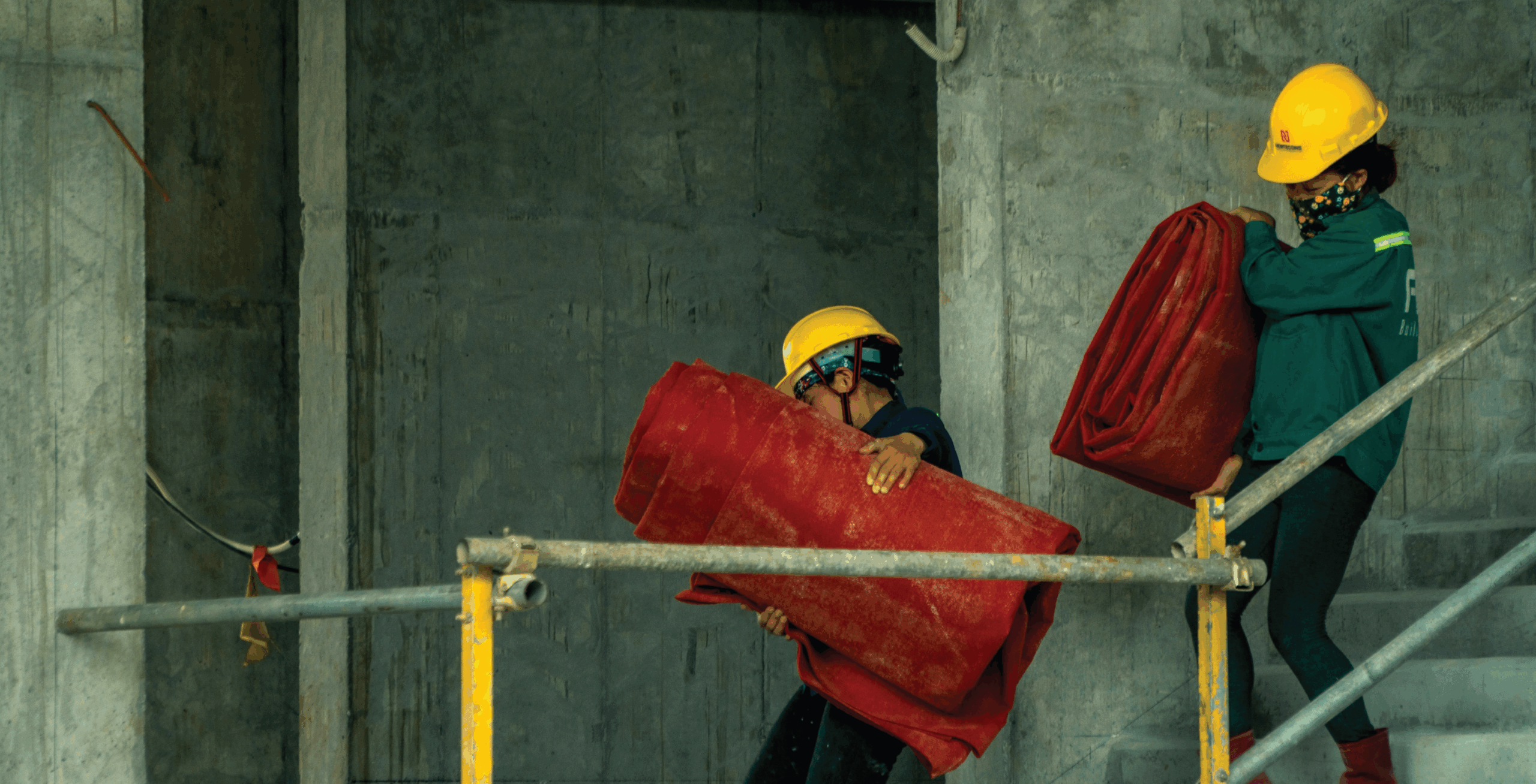 Two construction workers in yellow hard hats carrying large red rolls inside a concrete building under construction