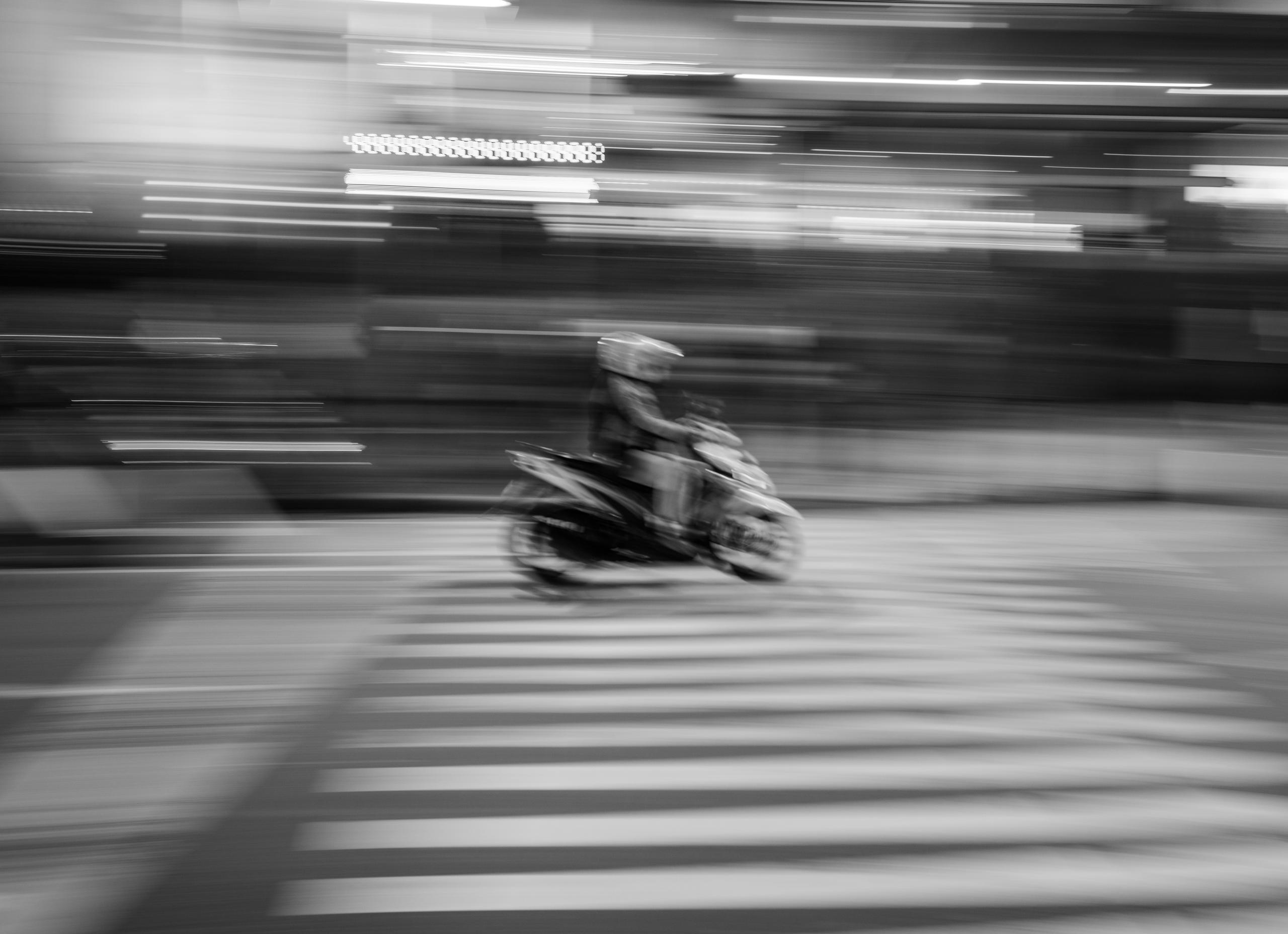 A blurred black-and-white scene of a motorcyclist speeding across a pedestrian crosswalk at night, with motion streaks from city lights emphasizing the rapid movement.