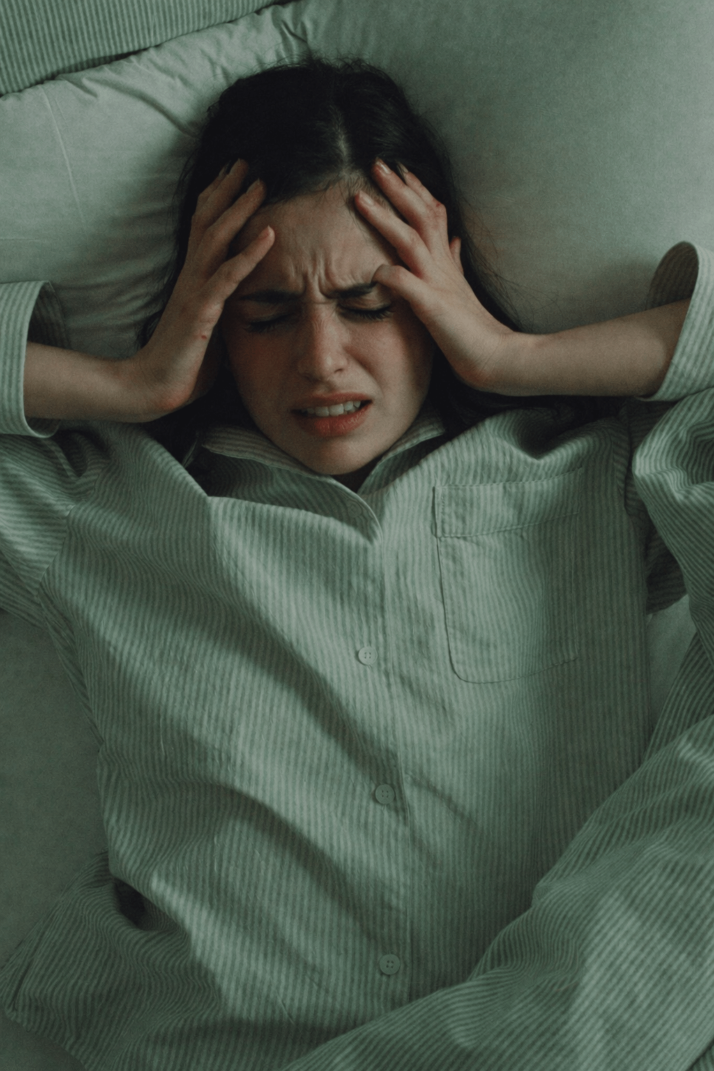 A woman lying in bed wearing green striped pajamas presses her fingers against her temples, her face tense and eyes squeezed shut as if experiencing a headache or pain.