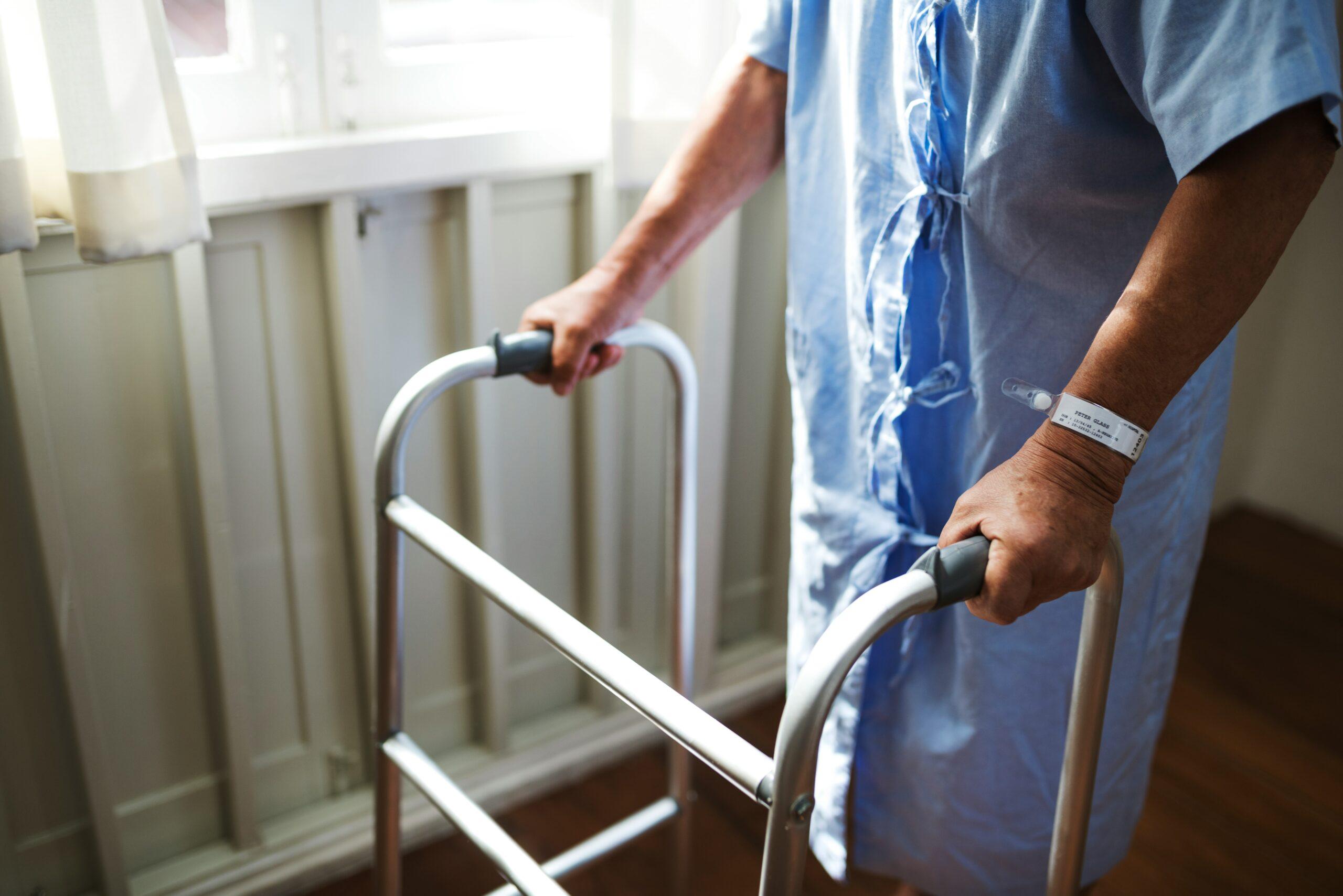 An older patient in a hospital gown gripping a metal walker with both hands, a medical ID band visible on one wrist, standing beside a window with soft daylight illuminating the room.