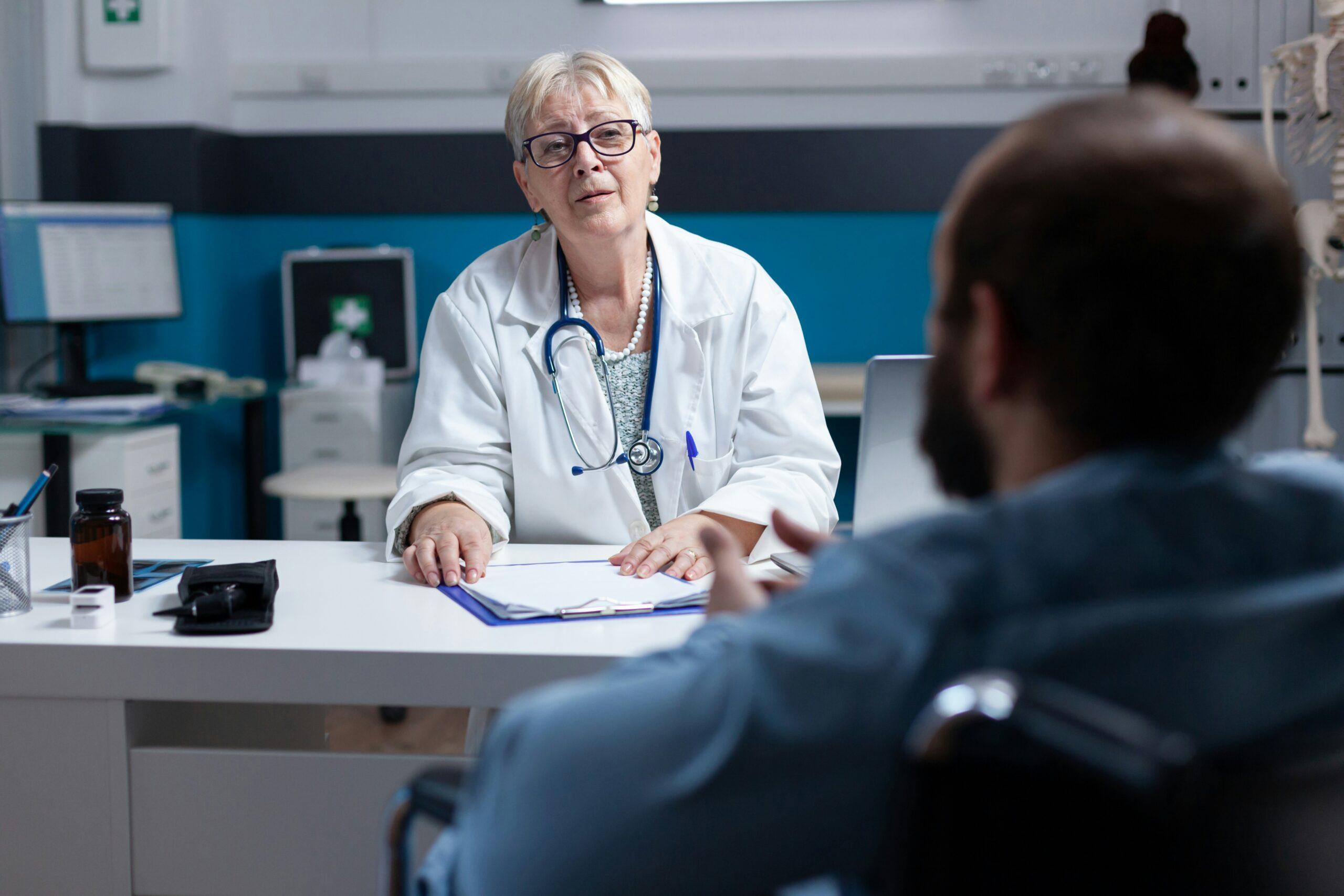 An older female doctor wearing a white lab coat and stethoscope sits at a desk in a medical office, listening attentively to a male patient seated across from her, with paperwork, a laptop, and medical equipment visible in the background.