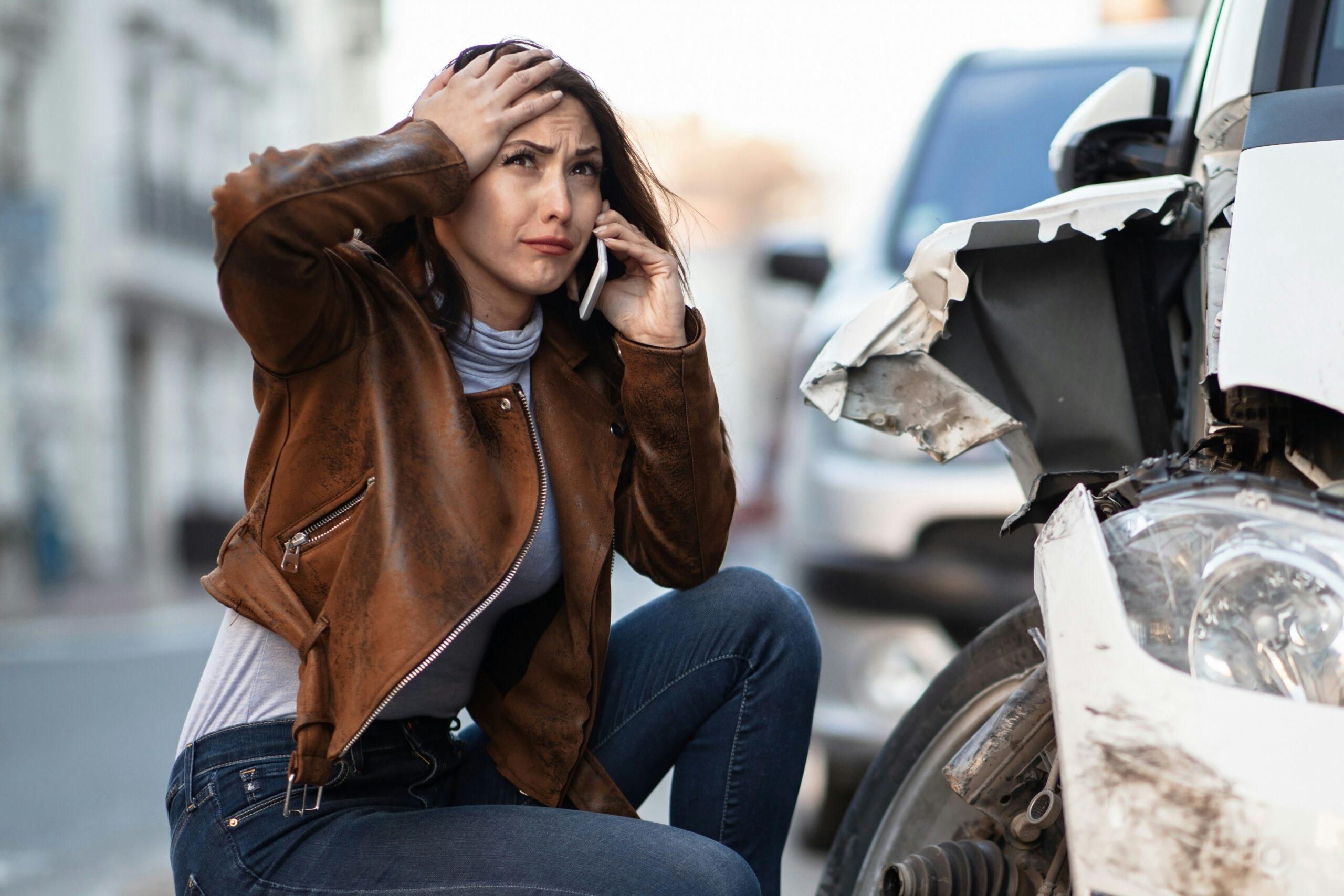 A distressed woman crouching beside a severely damaged car, holding her head with one hand while talking on the phone with the other, the crumpled front bumper and exposed internal parts of the vehicle visible in the foreground.