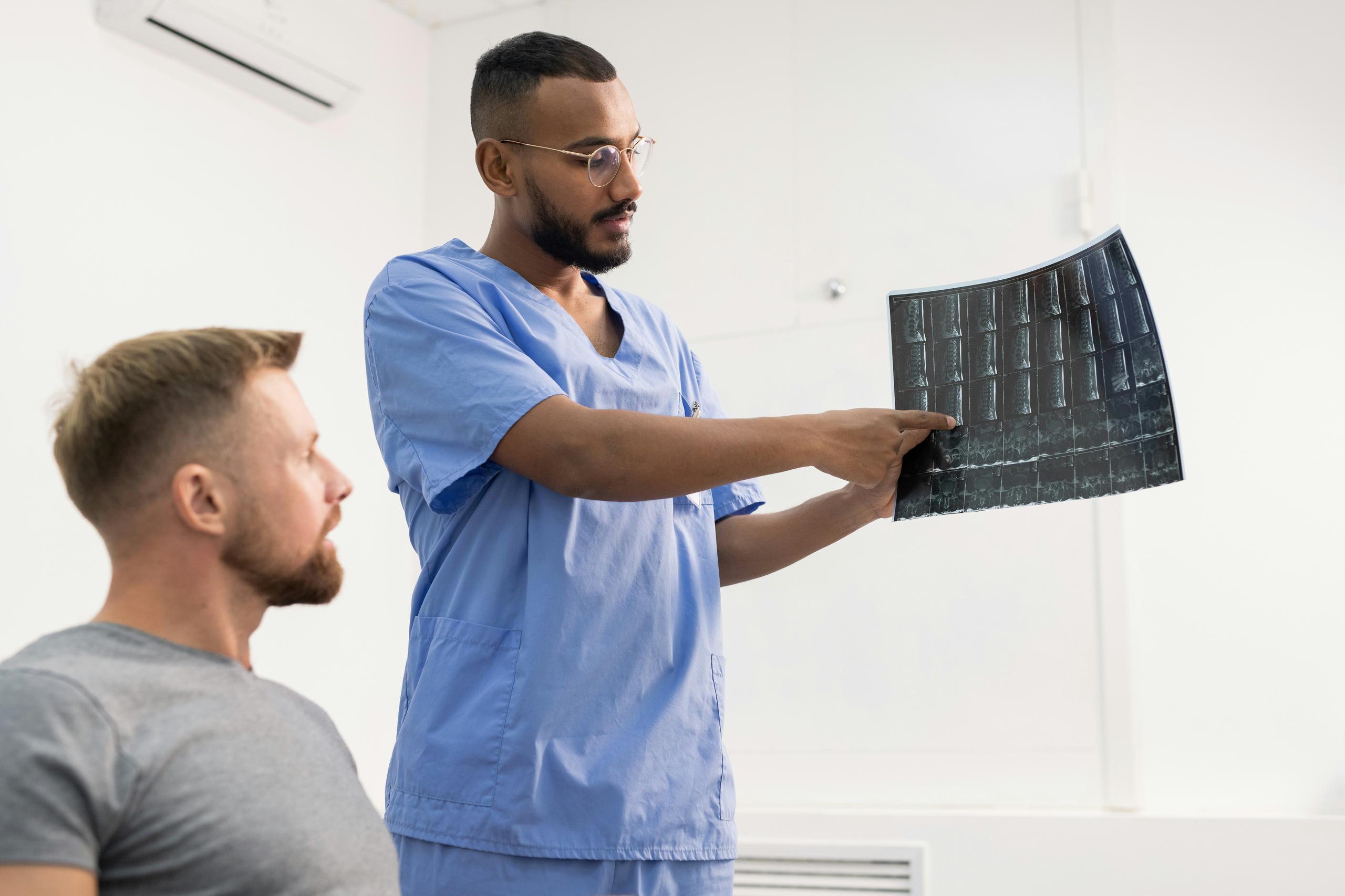 A healthcare professional in blue scrubs holding up and pointing to a spinal X-ray film while explaining the results to a seated patient in a clinical examination room.
