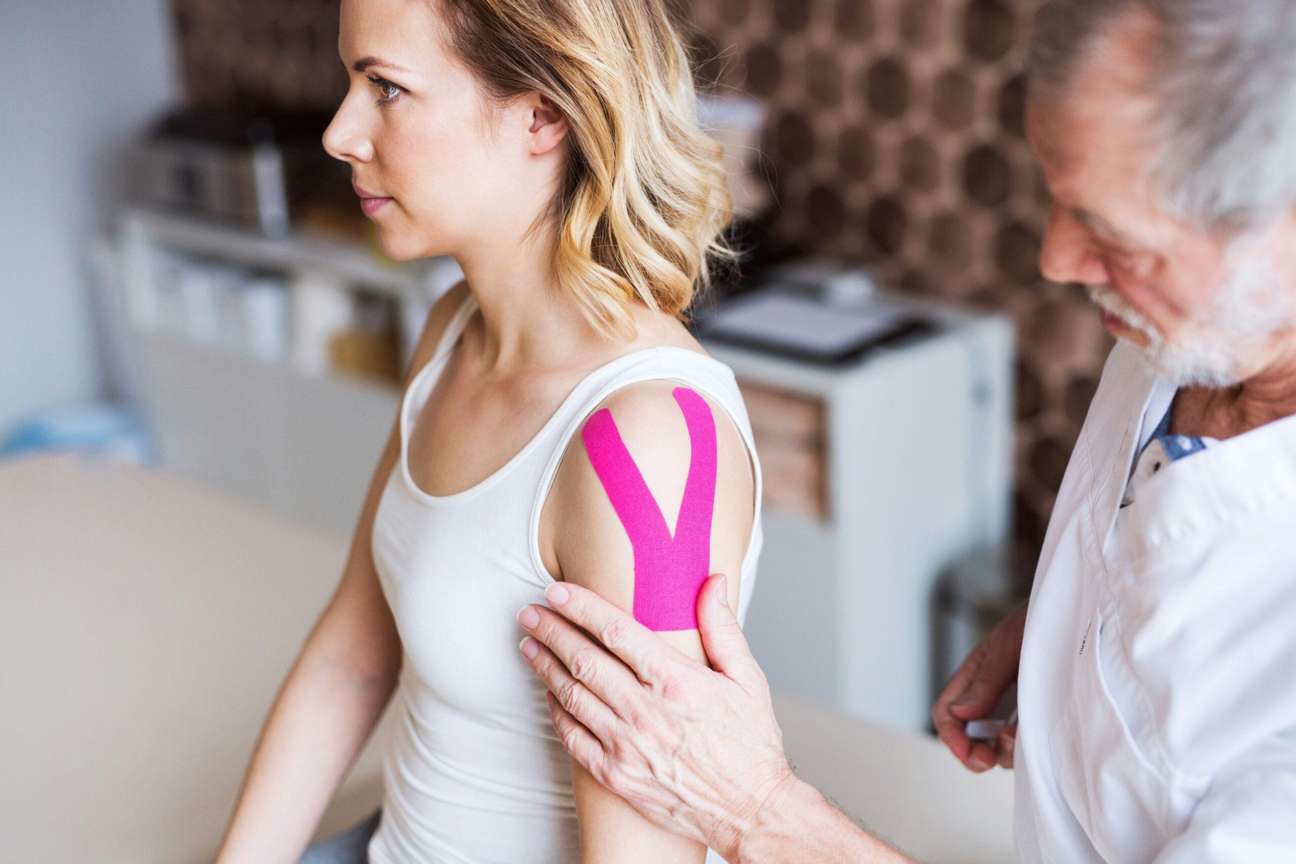 A woman sitting upright while a healthcare professional examines her shoulder, which is supported with bright pink kinesiology tape, in a clinical treatment setting.