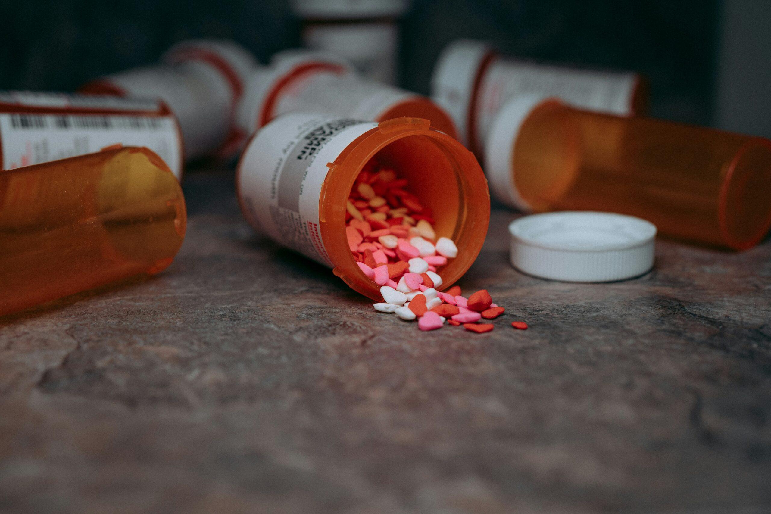 A spilled orange prescription bottle lying on its side with colorful heart-shaped candies scattered across a textured surface, surrounded by other empty pill containers in the background.
