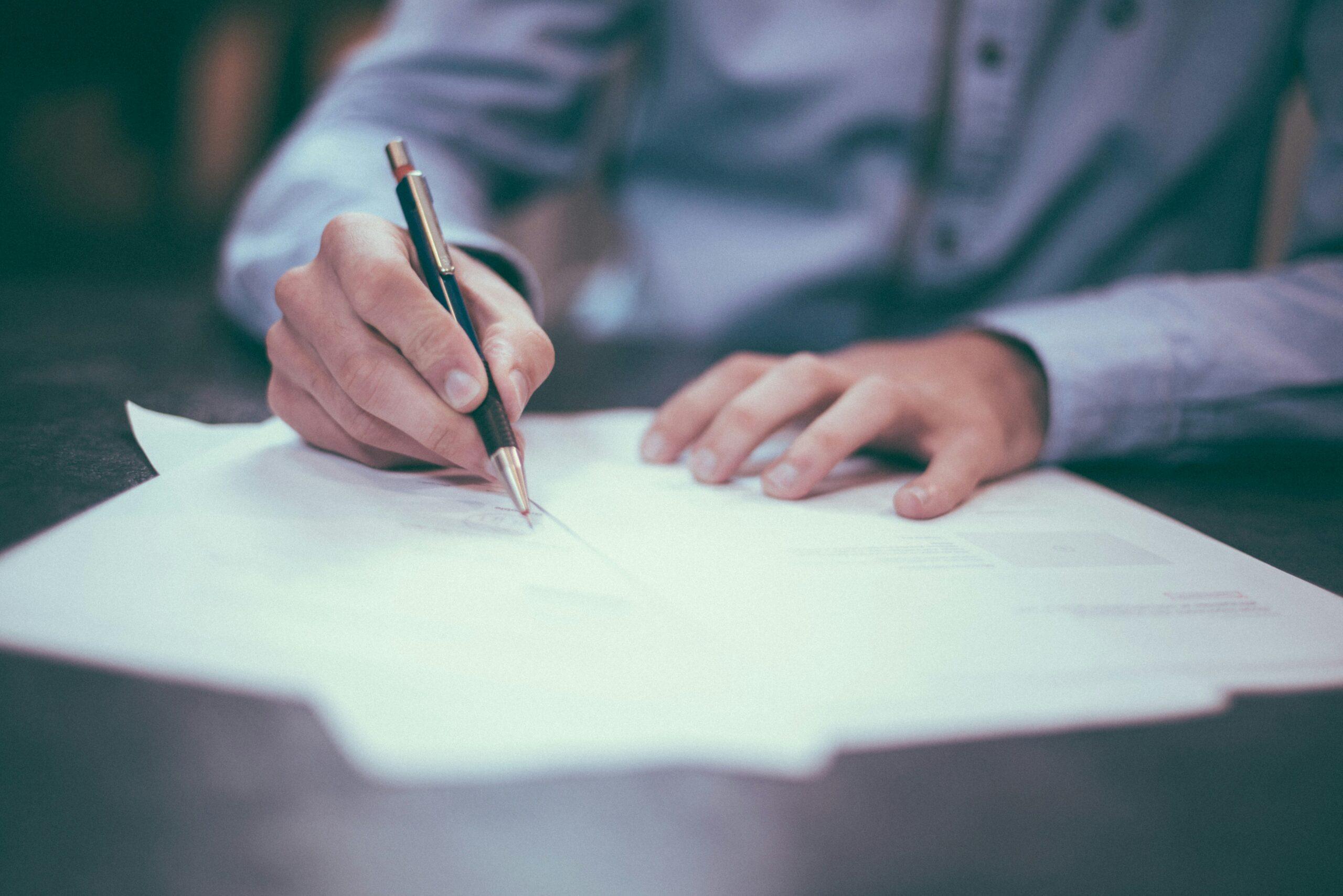 A close-up of a person in a button-down shirt writing on paperwork at a desk, with one hand holding a pen and the other steadying the documents.
