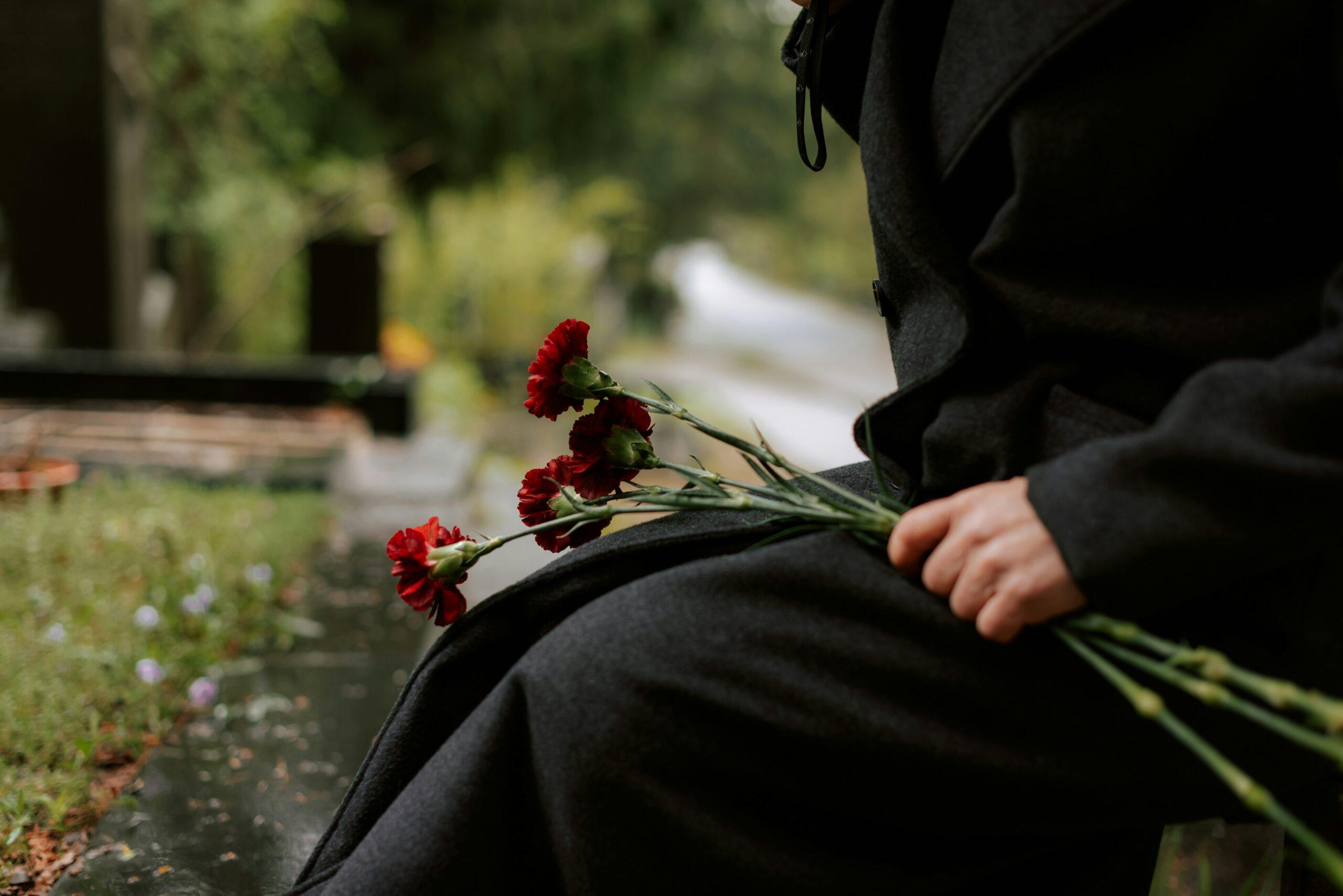 A person dressed in dark mourning clothes sitting beside a grave, gently holding a small bundle of red carnations with long green stems, surrounded by a soft, blurred background of greenery and headstones on a damp, overcast day.