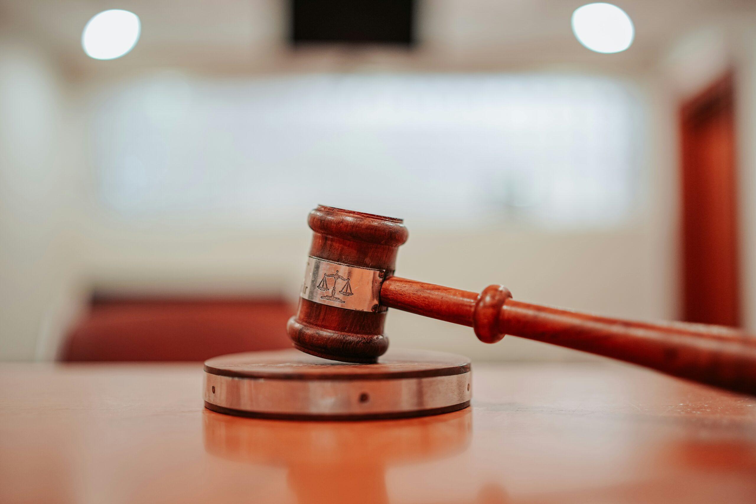 A close-up of a wooden judge’s gavel resting at an angle on its round sound block inside a bright courtroom, with soft blurred lights and warm wooden tones in the background.