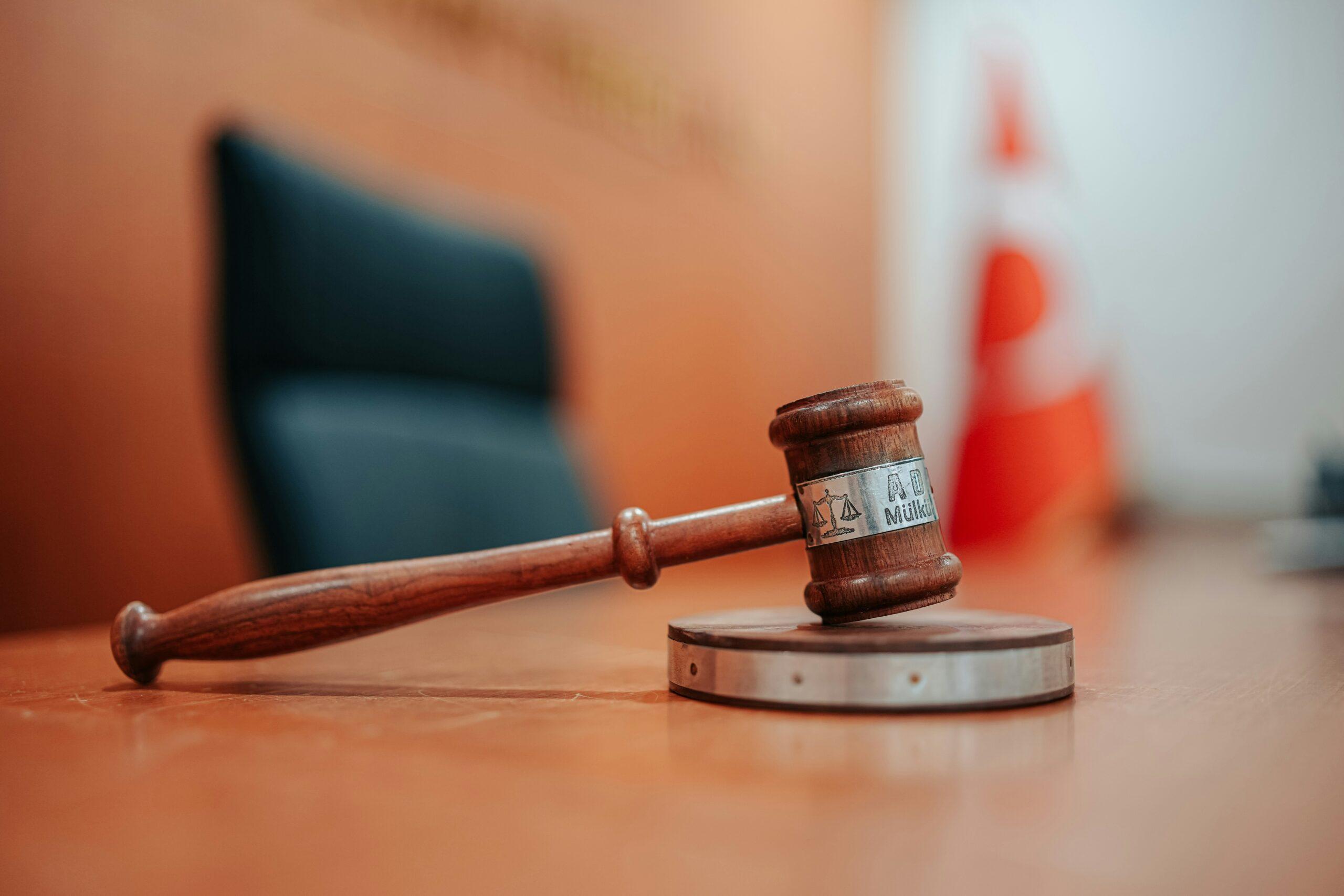 A wooden judge’s gavel resting on its sound block in an empty courtroom, with a blurred chair and a red-and-white object in the background creating a formal judicial setting.